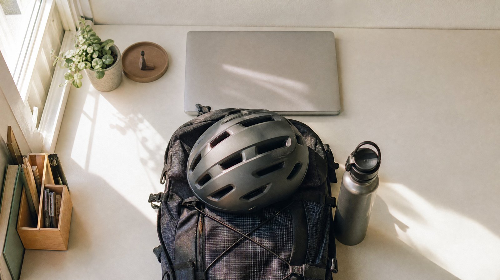 Von oben gesehen: ein gepackter Tagesrucksack mit Fahrradhelm liegt vor einem zugeklappten Laptop auf einem hellen Schreibtisch, daneben eine Trinkflasche, Mittagslicht fällt durchs Fenster