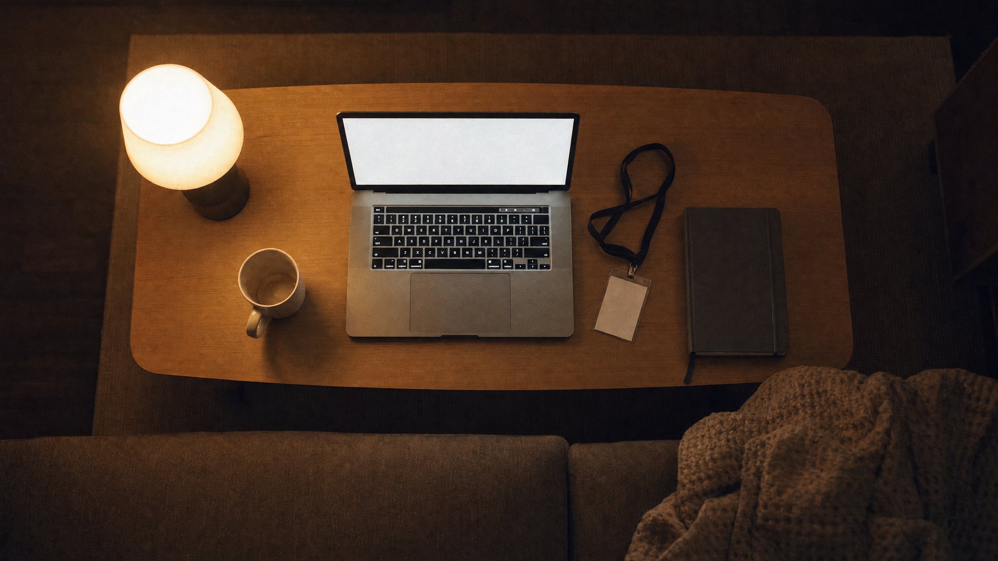 Von oben: ein aufgeklappter Laptop auf einem Wohnzimmertisch am Abend, daneben ein Firmenausweis an einem Schlüsselband, eine leere Tasse und ein geschlossenes Notizbuch, beleuchtet von einer warmen Tischlampe