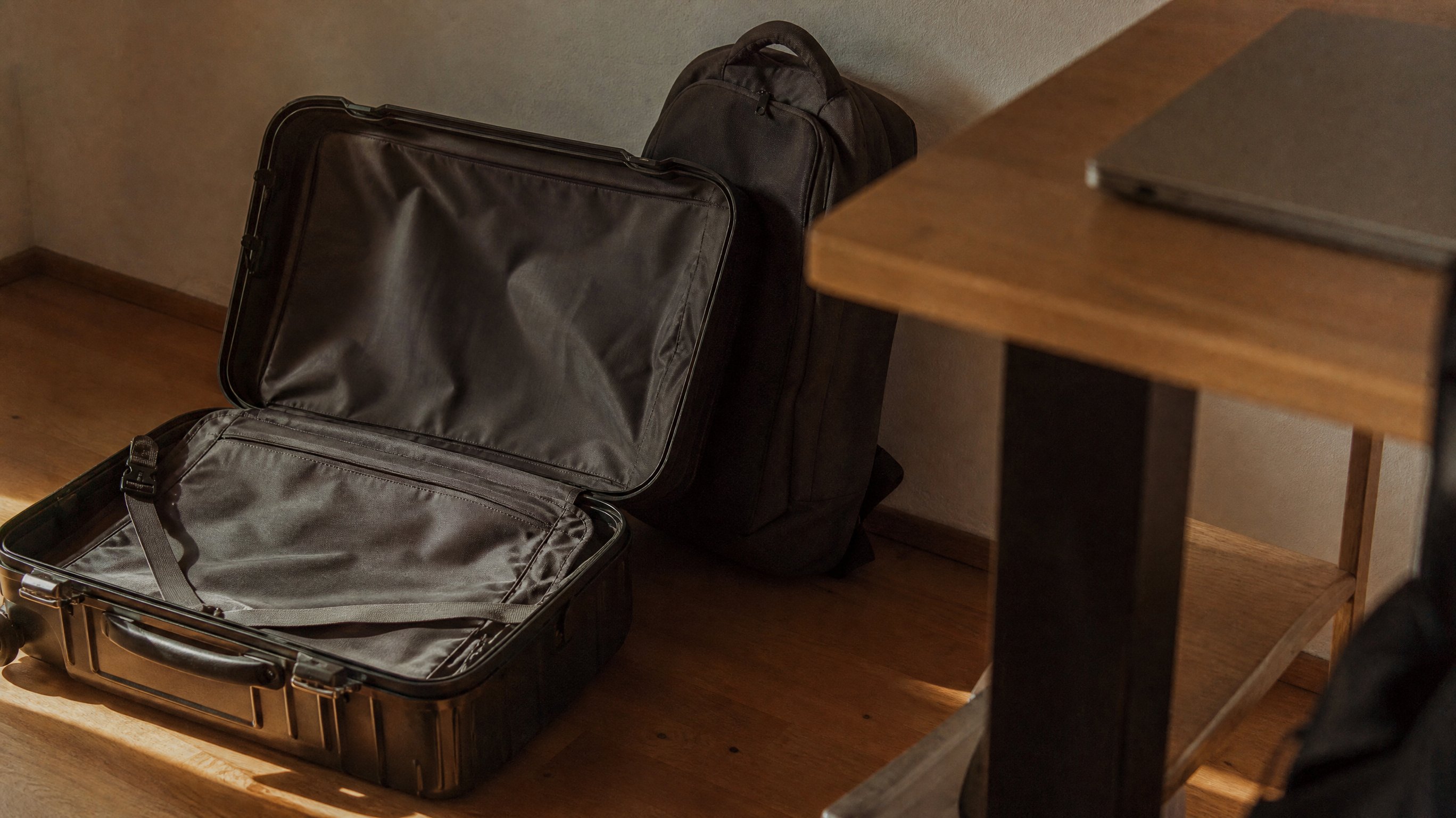 Ein offener, noch völlig leerer Kabinenkoffer auf dem Holzboden neben einem Schreibtisch, daran gelehnt ein Laptoprucksack, auf der Tischplatte darüber unscharf ein zugeklappter Laptop
