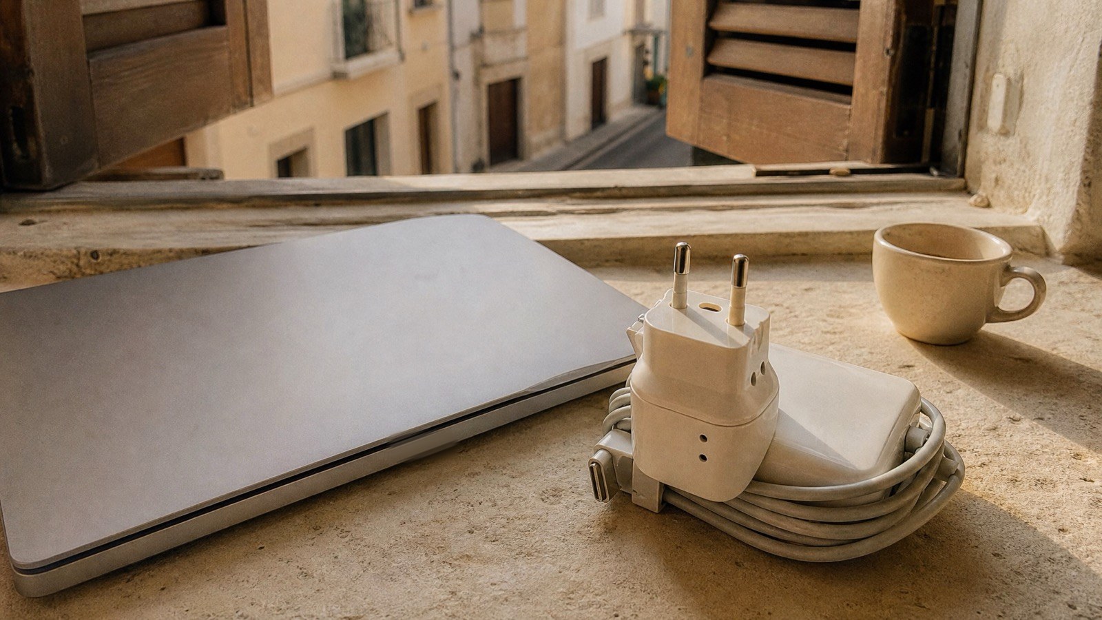Nahaufnahme auf einer breiten Fensterbank: ein Reise-Steckdosenadapter am aufgerollten Laptop-Netzteil neben einem zugeklappten Laptop, dahinter offene Holzläden und eine ruhige südliche Gasse im Morgenlicht
