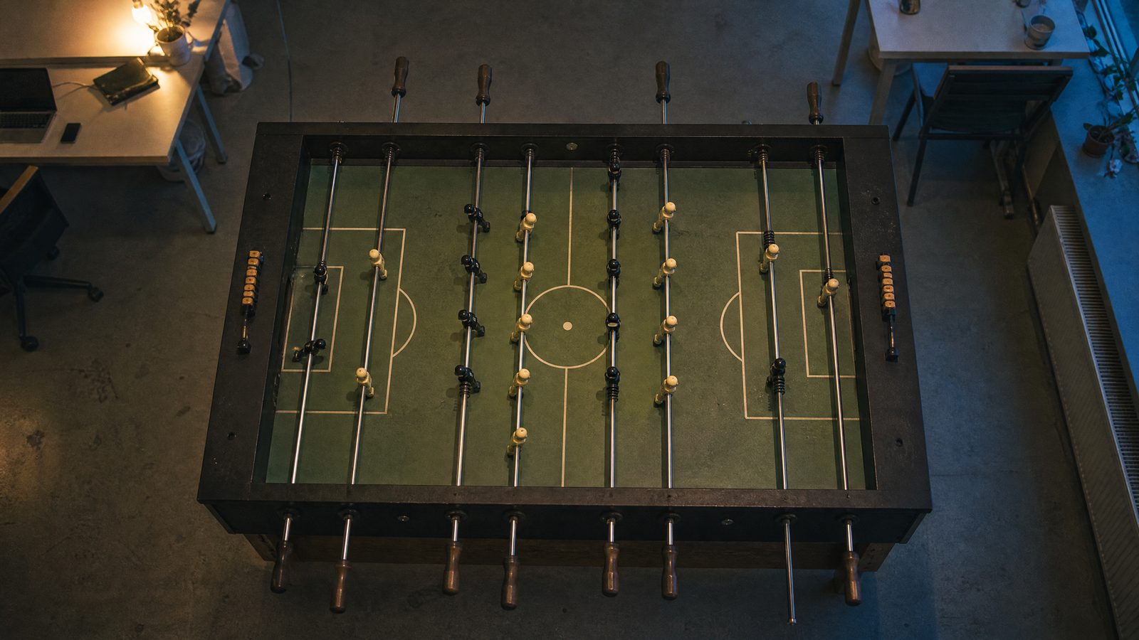 Blick von oben auf einen unbespielten Tischkicker in einem leeren Großraumbüro am frühen Abend, der Ball liegt am Anstoßpunkt, am Rand ein verlassener Schreibtisch mit Laptop im Licht einer Tischlampe und blaues Dämmerlicht vom Fenster